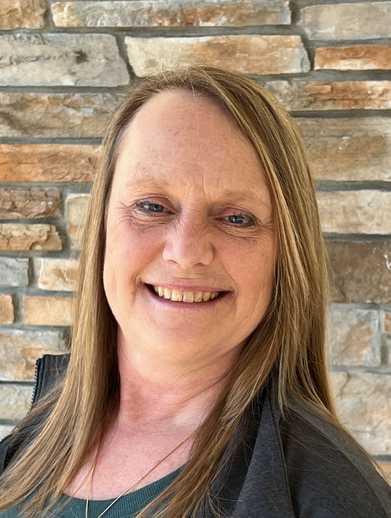 A woman with straight, light brown hair smiles at the camera. She is wearing a dark jacket over a green top, standing in front of a stone brick wall background.